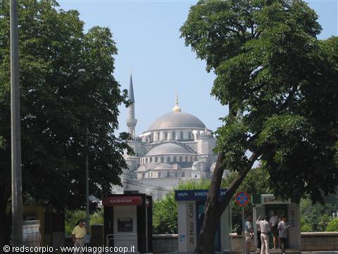 Istanbul's Blue Mosque behind green trees, kiosks and pedestrians under a clear sky