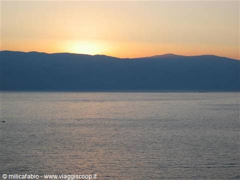 Dark mountains over calm sea, orange sky and low sun on the horizon