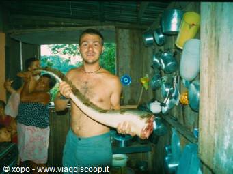 Shirtless man lifts a large fish in a wooden kitchen with hanging pots
