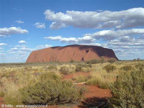 Uluru, red monolith surrounded by desert shrubs and sand under cloudy sky