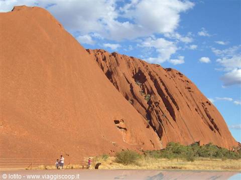 Tourists walk at the base of the red Uluru rock under a partly cloudy sky
