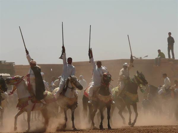 Horsemen in white robes raise rifles amid dust at a fantasia