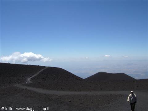 Hiker walks across dark volcanic craters on Mount Etna under blue sky