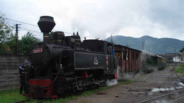 Black steam locomotive Cozia-1 releasing smoke, man beside, wet tracks, green mountains