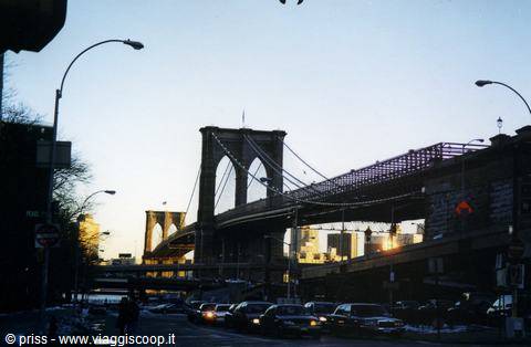 Brooklyn Bridge at dusk, traffic below, street lamps lit, pale blue sky