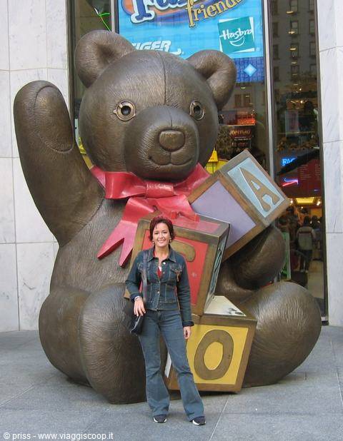 Woman in jeans beside a bronze teddy bear statue with colorful letter blocks outside a store