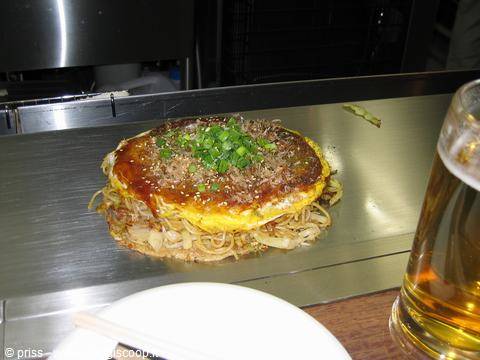 Okonomiyaki con uova, cipollotto e salsa sulla piastra d'acciaio, accanto a una birra