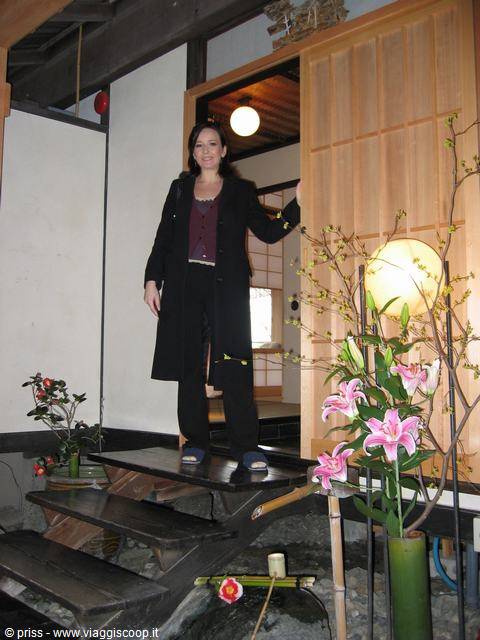 Donna in nero su gradini di legno all'ingresso di una casa tradizionale, fiori rosa