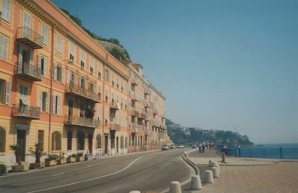 Ochre buildings line the waterfront, people walk by the blue sea