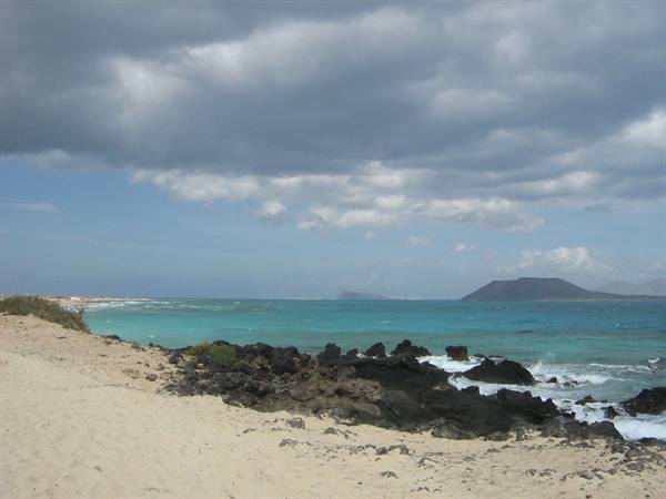 Pale sand and black rocks on turquoise sea, volcano behind, Corralejo