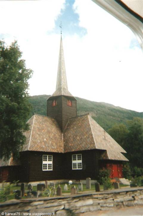 Old dark wooden stave church with tall spire, stone wall, graveyard, green mountains behind