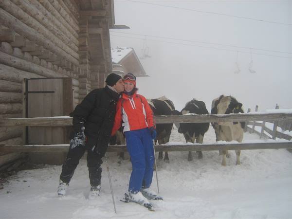 Skier kisses woman in red jacket by wooden cabin with cows behind fence