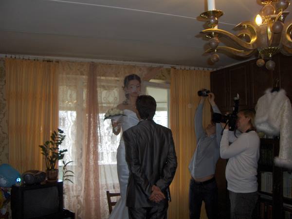 Bride in white gown with bouquet facing groom as photographers shoot, yellow curtains behind
