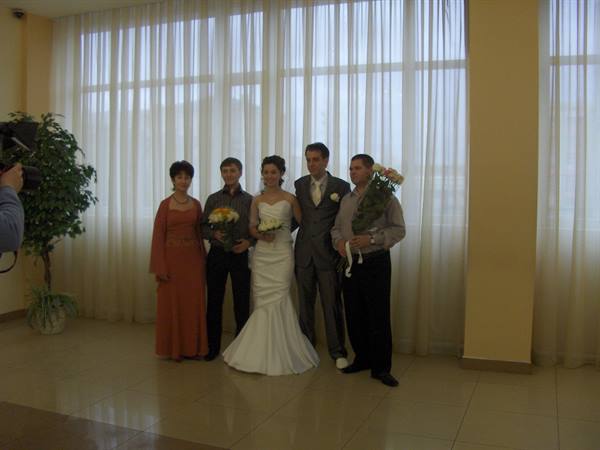 Bride in white gown and groom pose with guests holding flowers by sheer curtains