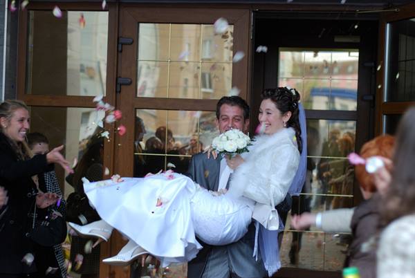 Groom in gray suit carries laughing bride in white among flying petals