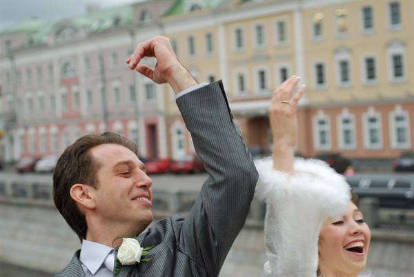 Groom with roses and laughing bride celebrating in front of pastel classical buildings