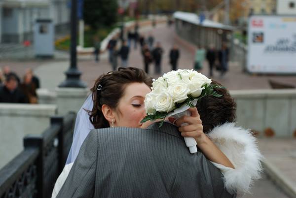 Bride with white rose bouquet embraces groom on a city bridge
