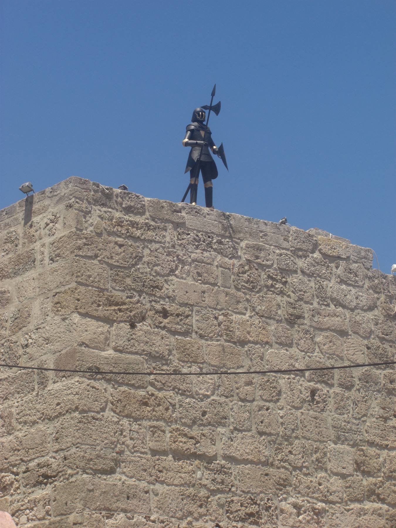 Knight statue in armor with axe atop Rhodes fortress walls, stone, sunlight