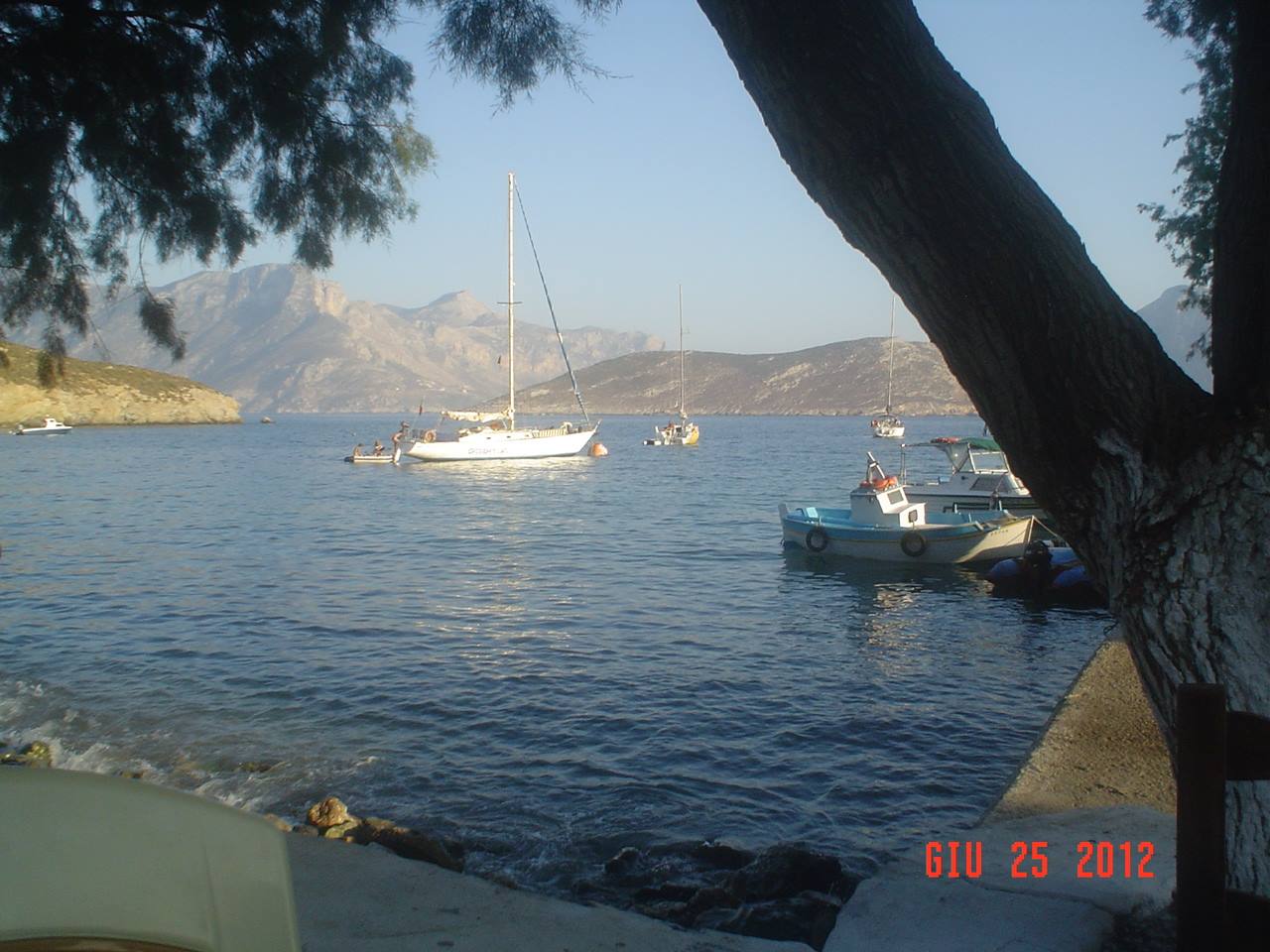 Boats anchored in calm bay beneath arid mountains, tree branches framing foreground