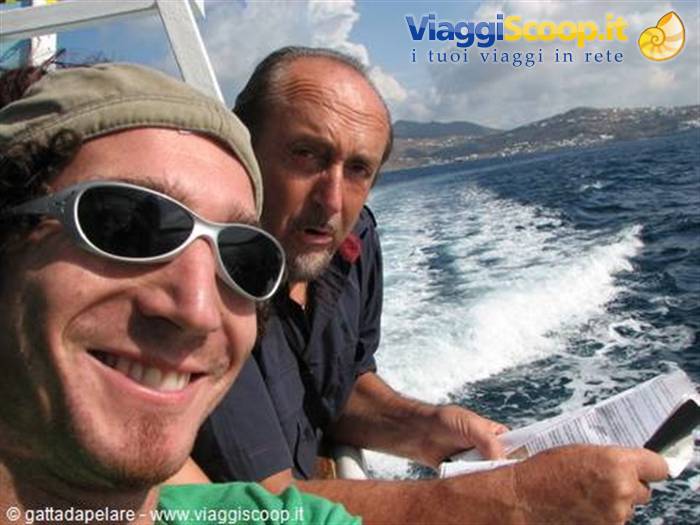 Two men on a ferry, white wake behind and coast with white houses