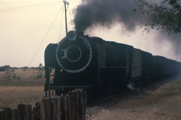 Black steam locomotive puffing dense smoke past dry fields and wooden fence