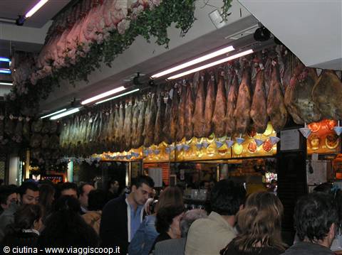 Cured hams hanging under fluorescent lights in a crowded deli counter