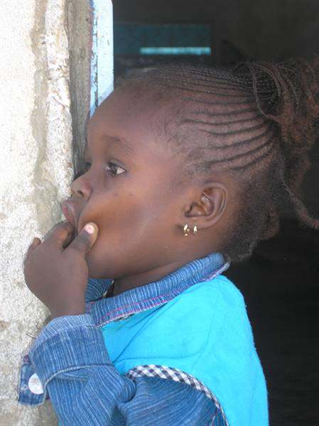 Little girl with braids and earring beside a white wall, hand at her mouth