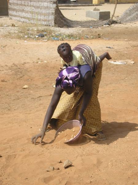 Woman carrying a child on her back sieves sand with a purple sieve