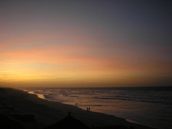 Orange and purple sky over the ocean, two people walk along the dark beach