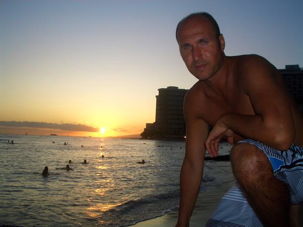 Man crouching on Waikiki sand, swimmers in the water at sunset