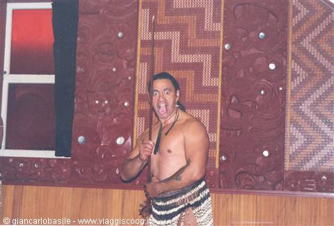 Maori man with spear grimacing before carved wooden meeting house