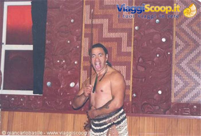 Maori man with spear grimacing before carved wooden meeting house