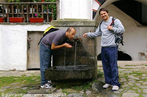 Tourist drinks from an old stone fountain while his friend smiles beside