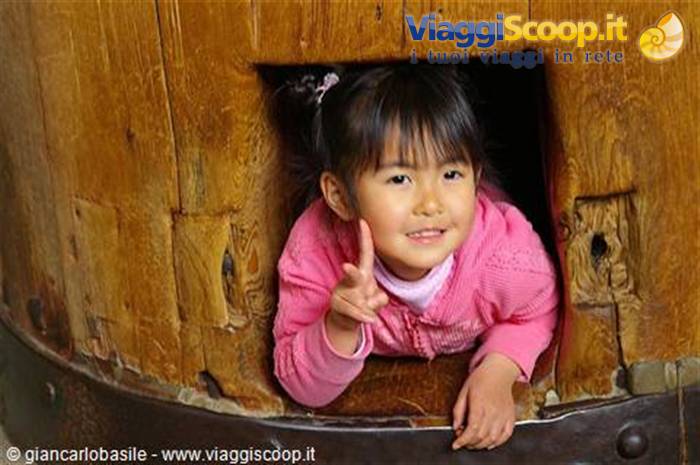 Little girl in pink jacket peeking out of wooden barrel, gesturing