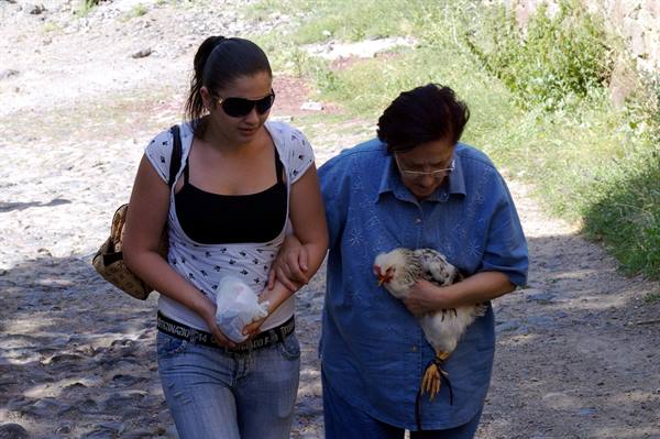 Two women walk on a dirt path, one cradling a chicken in her arms
