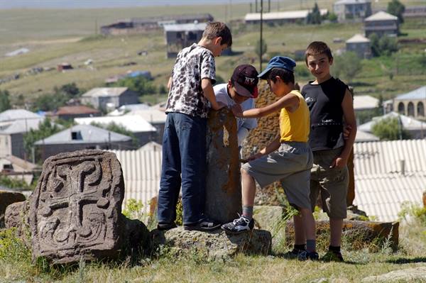 Boys play beside a carved stone khachkar, village houses on the hillside behind