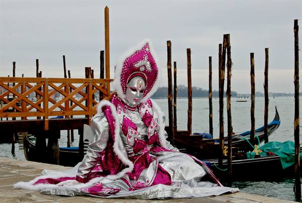 Venice carnival figure in silver and pink costume sitting on a quay beside gondolas