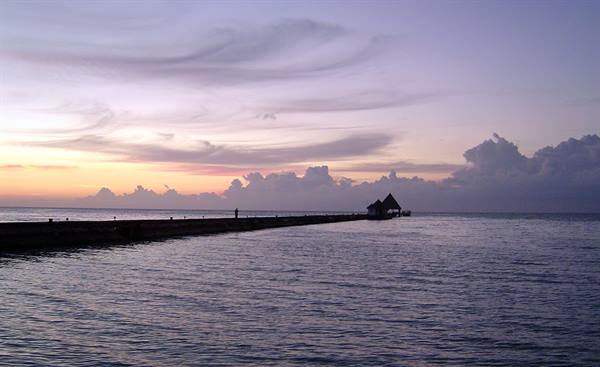 Wooden pier crossing calm sea toward small pavilion, purple and orange sky