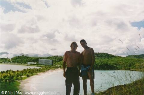 Two young men on a grassy hill above a turquoise bay with beach and hotel