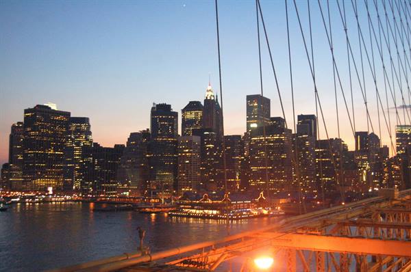 Manhattan skyscrapers lit at dusk, Brooklyn Bridge cables in the foreground