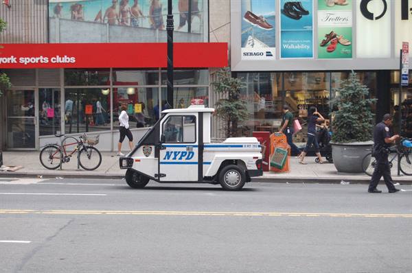 NYPD three-wheeler parked by midtown shops, officer walking past pedestrians