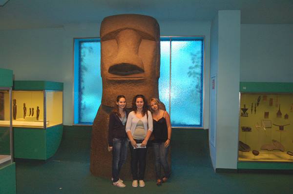 Three girls posing beside a moai statue in an ethnographic museum