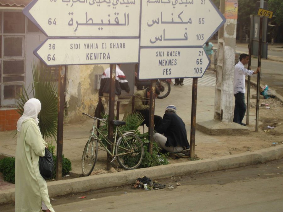 Woman in djellaba beside road signs pointing to Kenitra, Meknes and Fes