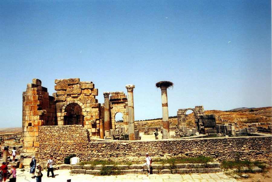 Roman ruins of Volubilis: stone columns and arches under blue sky, tourists walking