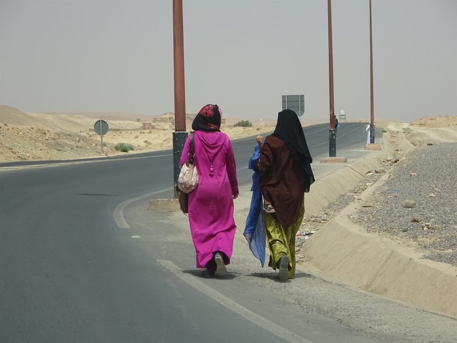 Women in djellabas walk along the asphalt road between sand and lampposts, Merzouga