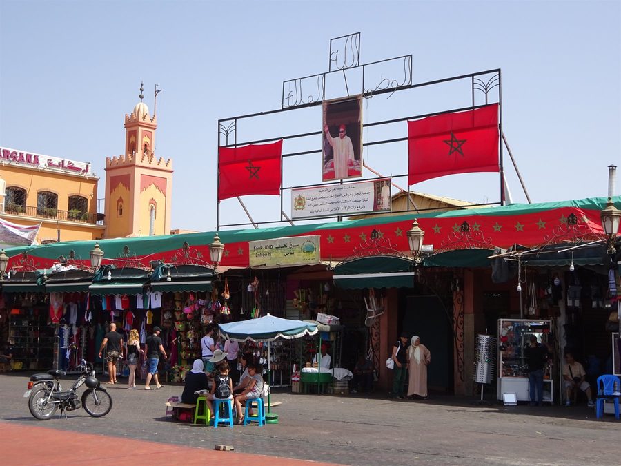 Jemaa el-Fna square in Marrakech: Moroccan flags, royal portrait, minaret and market stalls
