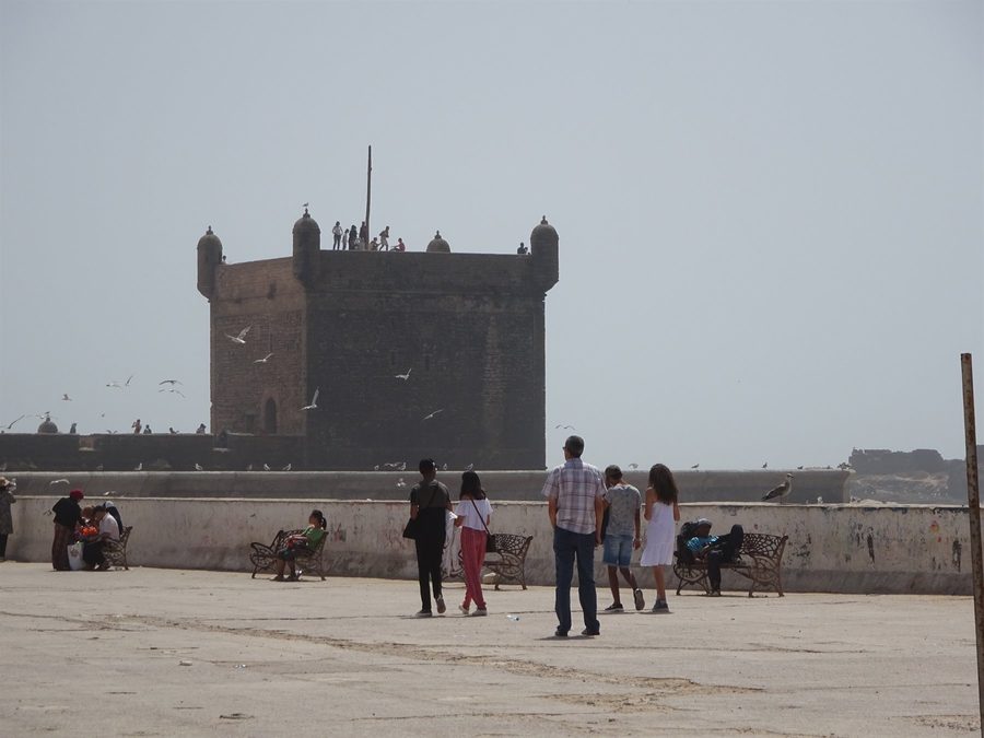 Tourists walk toward the Sqala du Port in Essaouira, seagulls in hazy sky